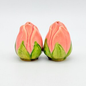 May include: A pair of ceramic salt and pepper shakers shaped like closed rosebuds. Each shaker features a glossy finish with soft pink petals at the top and vibrant green leaves at the base, with a single small hole at the very top of each.