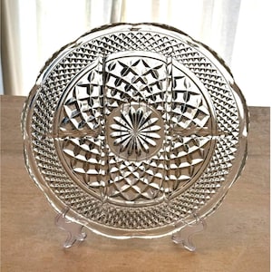 May include: A clear, round, divided serving platter with a decorative, cut-glass design. The platter features a central starburst pattern and is supported by a clear plastic stand. The rim has a scalloped edge, and the surface is divided into sections.