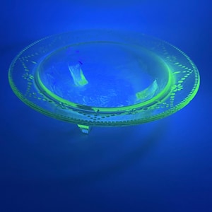 May include: A clear, round glass plate with a decorative floral and dot pattern around the rim. The plate has a slight green tint and is elevated by three small feet. The background is a deep blue.