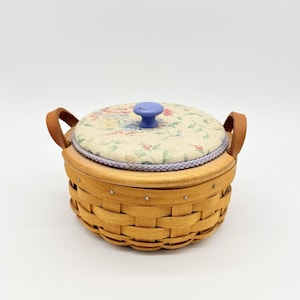 Op de afbeelding: Een ronde, geweven mand met een bloempatroon deksel en een blauwe knop. De mand heeft twee leren handvatten en een lichtbruine kleur. Het deksel is crèmekleurig met een bloemenpatroon in roze, blauw en groen.