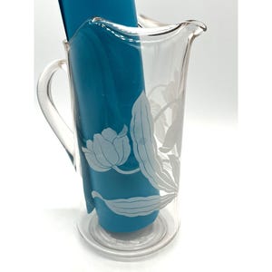 May include: Clear glass pitcher with a floral design. The pitcher has a handle and a spout for pouring. A teal-colored item is inside the pitcher. The floral design is etched into the glass, creating a delicate and elegant look. The pitcher is likely used for serving beverages.