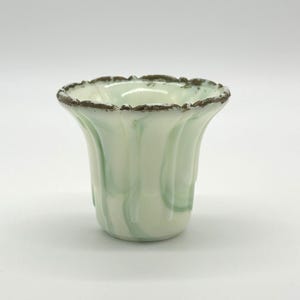 May include: A small, pale green and white glass vase with a ruffled rim and a flared shape. The vase has a swirled pattern of green and white, and the rim is trimmed with a dark brown color.