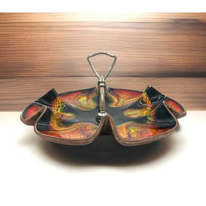 May include: A two-tiered serving dish with a black, red, and yellow marbled design. The dish has a silver handle and a scalloped edge. The dish is made of ceramic and is suitable for serving appetizers or desserts.