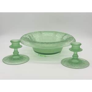 May include: A green glass centerpiece set featuring a round bowl and two matching candle holders. The bowl has a decorative rim and sits on small feet. The candle holders have a round base and a short, cylindrical top.