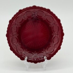 May include: A ruby red glass plate with a scalloped edge and embossed floral design. The plate has a textured center and is displayed on a clear plastic stand. The plate is a decorative piece.