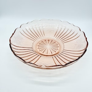 May include: A pink glass serving bowl with a scalloped edge. The bowl has a circular center with a starburst design and radiating lines. Floral patterns are etched around the rim. The bowl is likely vintage or antique.