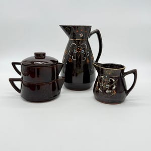May include: A dark brown ceramic tea set with a teapot, creamer, and pitcher. The set features floral designs in orange, blue, and white. The pitcher and creamer have gold trim.