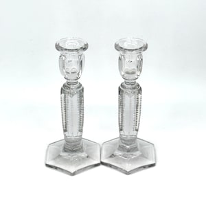 May include: A pair of clear glass candle holders featuring a hexagonal base and a vertical ribbed pattern on the central stem. Each holder has a circular top rim designed to support a candle, with a transparent finish and reflective surfaces.