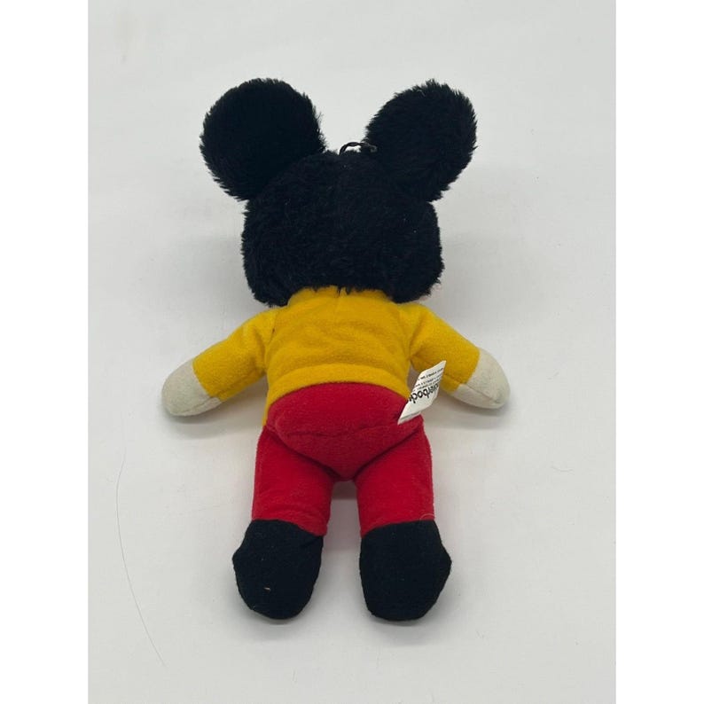 May include: A plush Mickey Mouse doll, viewed from the back, features a black head and ears, a yellow shirt, red pants, and black shoes. The doll has white hands and a small tag. This collectible toy is a classic.
