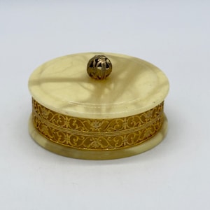 May include: A round, cream-colored stone trinket box with a polished finish. The sides are wrapped in a wide, intricate gold-tone metal filigree band. A small, spherical, open-work gold-tone metal knob sits centered on the flat lid.