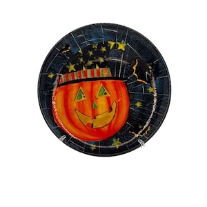 May include: A round Halloween plate with a smiling jack-o'-lantern wearing a striped hat. The orange pumpkin is set against a dark blue background with yellow stars and bats. The plate has a black rim, perfect for seasonal decor.