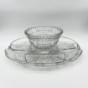 Puede incluir: Un juego de fuentes de vidrio transparente. El juego incluye un cuenco central, rodeado de seis platos más pequeños, todos conectados a una bandeja con borde festoneado. El vidrio tiene un patrón texturizado en forma de diamante.