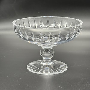 May include: A clear, footed glass bowl with a round base and a decorative, cut-glass design. The bowl has a wide, shallow shape with a scalloped rim and a pedestal base. The glass is transparent, allowing light to pass through.