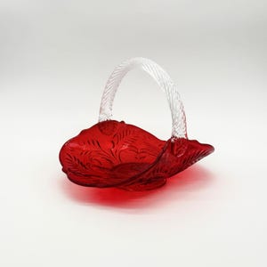Op de afbeelding: Een felrode glazen mand met een ingewikkeld reliëf bloemmotief op de kom en een helder, gedraaid glazen handvat. Het object heeft een geschulpte rand en een ronde voet, wat zorgt voor een doorschijnende afwerking door de gehele glasstructuur.