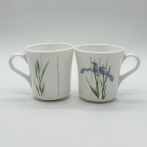 May include: Two white ceramic mugs with floral designs. One mug features a blue iris flower, while the other has a green stem and blue buds. Both mugs have a curved handle and are set against a white background.