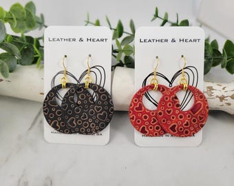 Puede incluir: Dos pares de pendientes colgantes con un diseño circular. Los pendientes están hechos de cuero y tienen un patrón de corazones y círculos. Los pendientes están en tarjetas blancas con el texto "Leather & Heart" y "Handmade in Atlanta, GA".