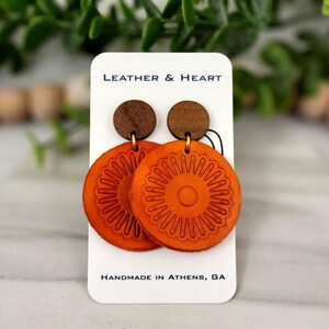 May include: A pair of orange leather earrings with a circular design and a brown wooden accent. The earrings are displayed on a white card with the text "Leather & Heart" and "Handmade in Athens, GA".