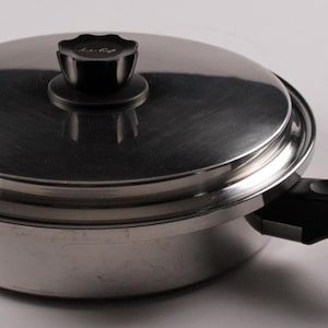 May include: Stainless steel skillet with a lid and black handle. The lid has a black knob. A pamphlet with green and white text is on the left side of the pan. The pan is made of stainless steel.