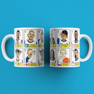May include: Two white ceramic mugs with a colorful cartoon design of soccer players. Each mug features a different set of players with their names and country flags. The mugs are on a blue background.