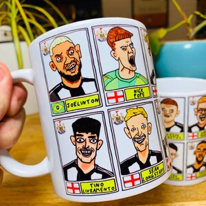 Puede incluir: Taza de cer&aacute;mica blanca con una ilustraci&oacute;n de dibujos animados de jugadores de f&uacute;tbol del Newcastle United. La taza presenta los nombres "Joelinton", "Nick Pope", "Tino Livramento" y "Sean Longstaff".