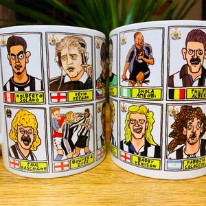 Peut inclure: Deux tasses en c&eacute;ramique blanche avec des poign&eacute;es noires. Chaque tasse pr&eacute;sente une grille de neuf illustrations de joueurs de football en dessin anim&eacute; avec leurs noms en dessous. Les tasses sont sur une surface en bois.