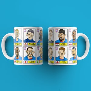 May include: Two white mugs with a blue and white design featuring cartoon illustrations of soccer players. The mugs have black outlines and yellow accents. The names of the players are printed below their images. The mugs are on a blue background.