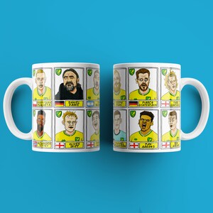以下が含まれることがあります： サッカー選手の漫画イラストが描かれた、黄色と緑のデザインの白いマグカップ2つ。マグカップには、画像の下に選手の姓名が印刷されています。マグカップのテキストには、「Teemu Pukki」、「Daniel Farke」、「Marco Stiepermann」、「Timm Klose」、「Oliver Skipp」、「Max Aarons」、「Alexander Tettey」、「Christoph Zimmermann」、「Grant Hanley」、「Todd Cantwell」、「Emi Buendia」が含まれています。