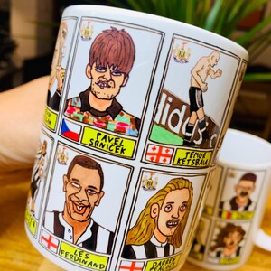 Peut inclure: Une tasse en c&eacute;ramique blanche avec une illustration de joueurs de football en dessin anim&eacute;. La tasse pr&eacute;sente un motif ray&eacute; noir et blanc avec le logo de Newcastle United. Les joueurs sont repr&eacute;sent&eacute;s dans un style humoristique avec des traits exag&eacute;r&eacute;s. La tasse porte les inscriptions "Les Ferdinand" et "Pavel Srnicek".