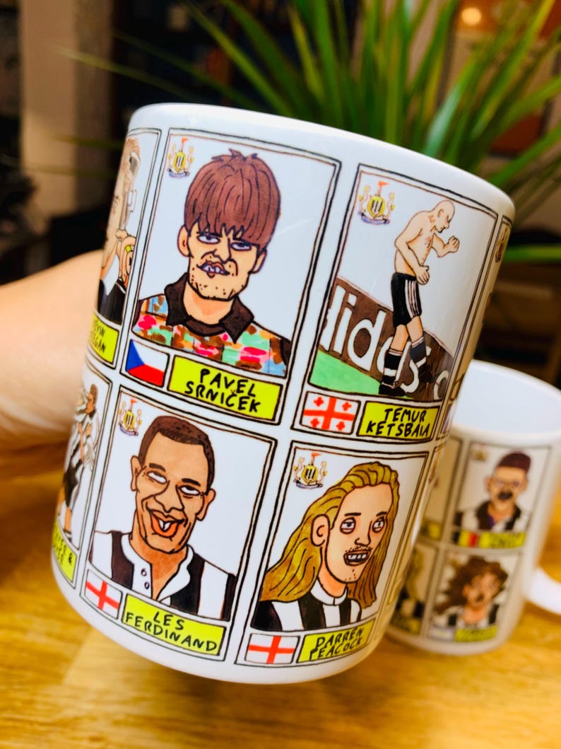 Peut inclure: Une tasse en c&eacute;ramique blanche avec une illustration de joueurs de football en dessin anim&eacute;. La tasse pr&eacute;sente un motif ray&eacute; noir et blanc avec le logo de Newcastle United. Les joueurs sont repr&eacute;sent&eacute;s dans un style humoristique avec des traits exag&eacute;r&eacute;s. La tasse porte les inscriptions "Les Ferdinand" et "Pavel Srnicek".