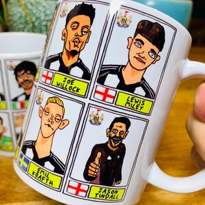 Puede incluir: Taza de cer&aacute;mica blanca con una ilustraci&oacute;n de dibujos animados de cuatro jugadores de f&uacute;tbol. La taza presenta un dise&ntilde;o de camiseta de f&uacute;tbol a rayas blancas y negras con detalles amarillos. Los nombres de los jugadores est&aacute;n impresos debajo de sus im&aacute;genes: Joe Willock, Lewis Miley, Emil Krafth y Jason Tindall.