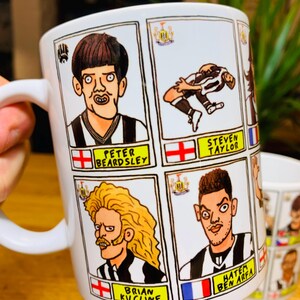 Peut inclure: Une tasse en c&eacute;ramique blanche avec des illustrations de joueurs de football en dessin anim&eacute;. Chaque joueur a un nom et un drapeau repr&eacute;sentant son pays d'origine. Les joueurs incluent Peter Beardsley, Steven Taylor, Brian Kilcline et Hatem Ben Arfa.
