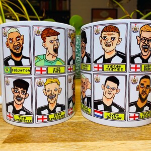 Puede incluir: Dos tazas de cer&aacute;mica blanca con asas negras. Cada taza presenta una cuadr&iacute;cula de ilustraciones de jugadores de f&uacute;tbol de dibujos animados con sus nombres y los logotipos de sus equipos. Las tazas est&aacute;n sobre una superficie de madera.