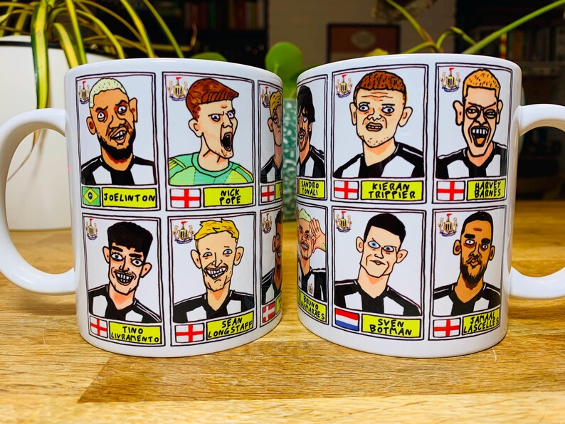 Puede incluir: Dos tazas de cer&aacute;mica blanca con asas negras. Cada taza presenta una cuadr&iacute;cula de ilustraciones de jugadores de f&uacute;tbol de dibujos animados con sus nombres y los logotipos de sus equipos. Las tazas est&aacute;n sobre una superficie de madera.