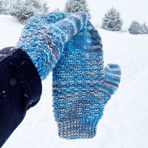 Puede incluir: Una manopla de punto azul y gris con un patrón de rayas blancas. La manopla se sostiene contra un fondo nevado.