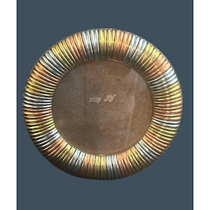 May include: A round picture frame featuring a wide, textured border composed of alternating segments in silver, gold, and copper tones. The center of the frame contains a plain, light brown backing material with a small, light-colored adhesive residue.
