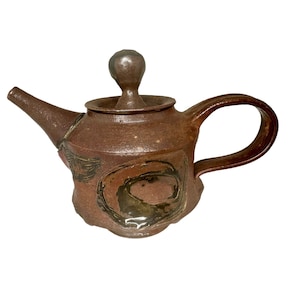 May include: A brown ceramic teapot with a rounded body and a lid with a knob. The teapot has a brown glaze with black and brown abstract designs.