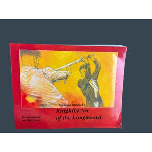 May include: A book titled "Knightly Art of the Longsword" by Sigmund Ringeck, with illustrations of sword fighting. The cover is red with a yellow background behind the illustrations. The book also lists David Lindholm and Peter Svärd as authors.