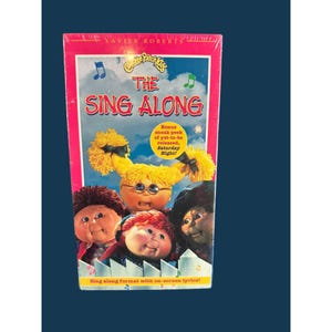 May include: A Cabbage Patch Kids The Sing Along VHS tape featuring a group of dolls with yarn hair and round faces. The cover displays the title in red text, blue musical notes, a yellow sunburst graphic, and a white picket fence illustration.