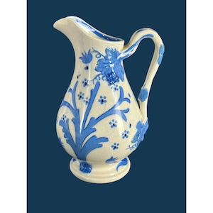 Peut inclure: Un pichet en céramique de couleur crème avec une finition brillante et une texture finement craquelée. Le récipient est orné de motifs floraux, de vignes feuillues et de grappes de raisin peints en bleu. Il possède une anse courbée.