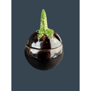 May include: A ceramic eggplant-shaped container with a lid. The container is a deep, glossy purple with a green stem and cap. The lid fits snugly, suggesting it's designed for storage or display.