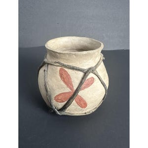 May include: A small, round, beige clay pot with a red flower design. The pot is wrapped with dark brown twine.