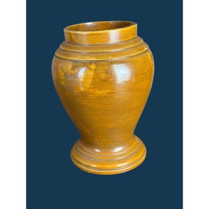 May include: A mustard-yellow ceramic vase with a smooth, glossy finish. The vessel features a wide, rounded body that tapers toward a narrow neck and a flared rim. It stands on a circular, tiered pedestal base with visible horizontal ridges.