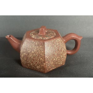 Könnte beinhalten: Eine rote Tonteekanne mit geometrischem Design. Die Teekanne hat einen Deckel und einen Griff. Die Teekanne ist mit einem wirbelnden Muster verziert.