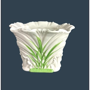 Puede incluir: Jarrón de cerámica blanca con la parte superior con volantes y cuerpo acampanado. El jarrón presenta diseños verdes pintados en forma de hierba en la parte delantera y una base verde. El jarrón es probablemente para fines decorativos.