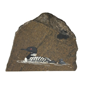 Pode incluir: Uma obra de arte pintada à mão em uma pedra retrata um mergulhão e seu filhote nadando na água. O mergulhão tem cabeça preta, dorso salpicado de branco e olho vermelho. A pedra tem formato irregular e cor marrom.