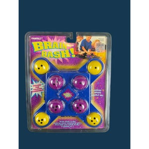May include: A Brain Bash electronic game in original blister packaging. The blue plastic console features four purple buttons and four yellow buttons. The packaging includes the text Brain Bash, Follow the Talking Command, and Great Music Built In.