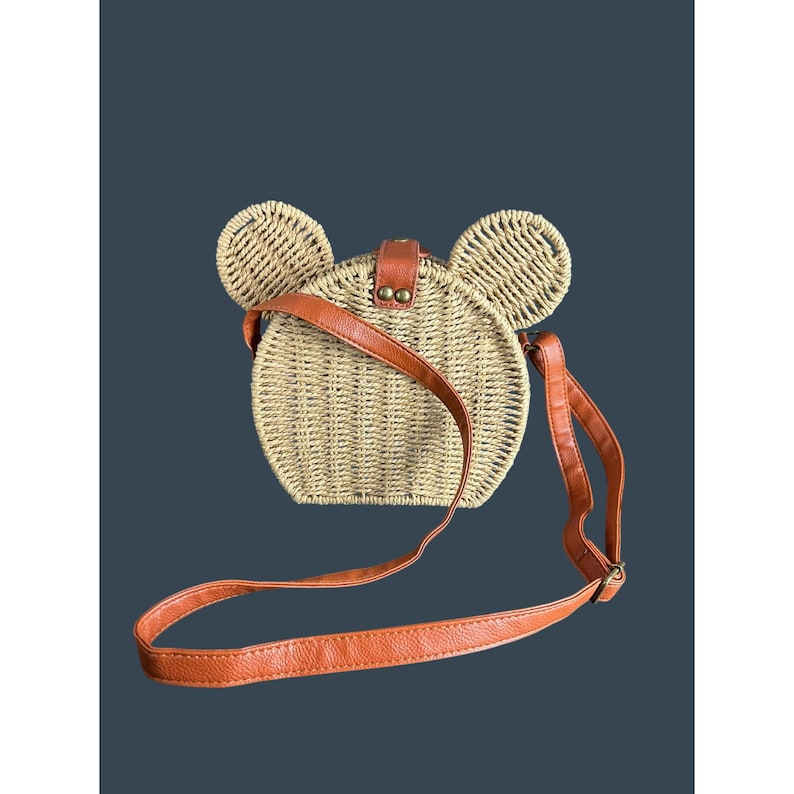 May include: A round, woven straw handbag with two large, rounded ears on top, resembling a mouse. The bag features a brown leather strap and a matching closure. The bag is tan and brown.