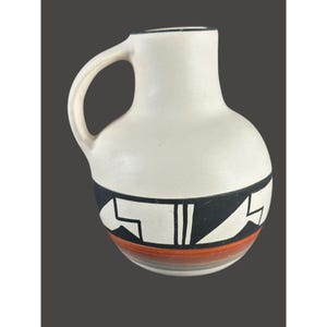 May include: A ceramic pitcher with a rounded body and a single handle. The upper portion is matte white, while the lower section features a wide black band with geometric white patterns, followed by a horizontal orange stripe and a grey base.