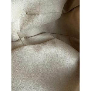 May include: Close-up of a beige fabric with visible folds and seams. The fabric appears to be a natural material, possibly linen or cotton, with a slightly textured surface. The lighting creates shadows that emphasize the fabric's texture and depth.