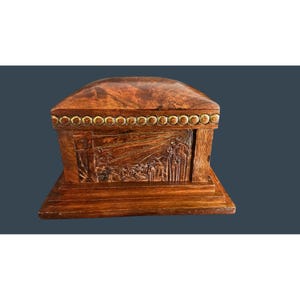 Puede incluir: Una caja de madera con tapa con bisagras. La caja es de color marrón cálido con una escena de paisaje tallada en la parte delantera. La tapa es redondeada y tiene un ribete decorativo de color dorado. La caja se asienta sobre una base rectangular.